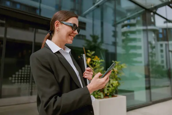 Stock image A professional woman in stylish glasses uses her smartphone in front of a contemporary office building