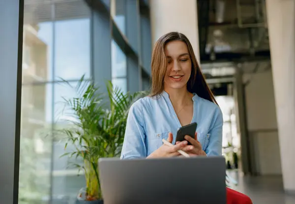 stock image A woman with a blue shirt using a smartphone and laptop while sitting in a bright modern office space