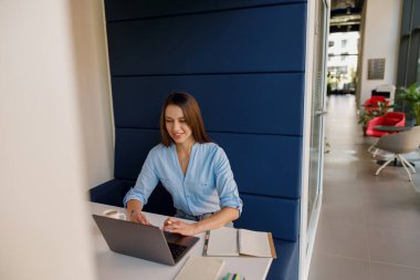 Mavi gömlekli iş kadını, bir ofis kabinindeki dizüstü bilgisayarda, elinde kalem ve defterle özenle çalışıyor..