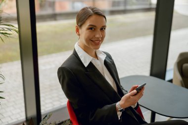 Resmi giyinmiş kendine güvenen bir iş kadını modern ofis ortamında gülümsüyor ve elinde bir akıllı telefon tutuyor.