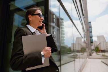 Ofis binasının dışında dizüstü bilgisayar tutarken akıllı telefon kullanan resmi takım elbiseli kendine güvenen bir iş kadını.