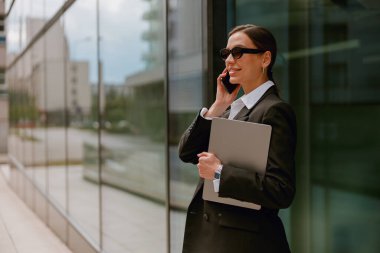 Güneş gözlüklü, modern ofis binasının dışında akıllı telefon kullanan, elinde dizüstü bilgisayar tutan kendine güvenen bir iş kadını..