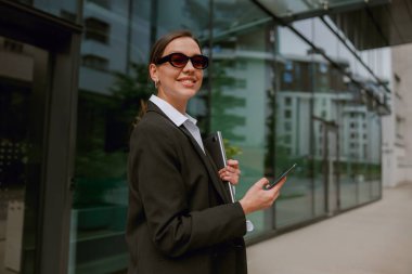 İş kıyafeti giymiş profesyonel bir kadın, modern bir ofis binasının önünde dururken akıllı telefon kullanıyor.