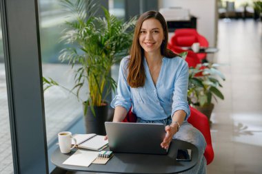 Mavi tişörtlü gülümseyen kadın dizüstü bilgisayarda çalışıyor, modern ofis salonunda rahatça oturuyor..