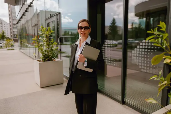 stock image Confident businesswoman in stylish suit with laptop and pen standing outside modern office building.