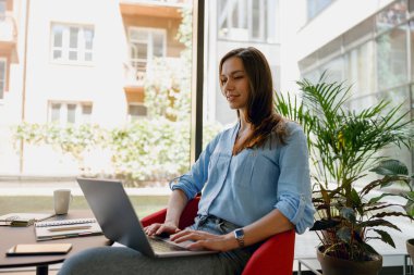 Genç bir profesyonel, modern bir ofis alanında dizüstü bilgisayarla çalışır. Doğal ışık ve bitkilerle..