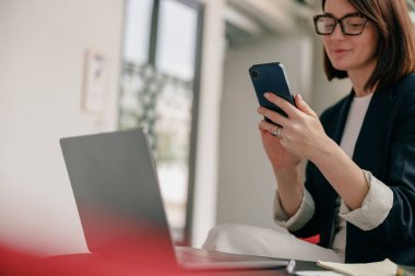 Modern bir ofiste akıllı telefon ve dizüstü bilgisayarı olan iş kadını çoklu görevleri, dinamik bir çalışma ortamı gösteriyor