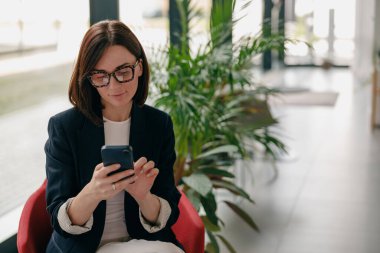 Ofis kıyafetleri içinde yeşil bitkiler ve doğal ışıkla çevrili akıllı telefon kullanan profesyonel bir iş kadını.