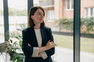 Modern ofis ortamında duran kendine güvenen bir iş kadını, kollarını kavuşturup pencereden dışarı bakıyor..