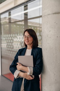 Ofisteki profesyonel kadın dizüstü bilgisayarla modern ortamda duruyor, kendine güveni tam..