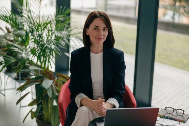 Modern ofiste oturan kendine güveni tam bir iş kadını. Etrafı bitkilerle ve doğal ışıklarla çevrili bir dizüstü bilgisayarla çalışıyor..