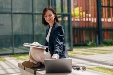 Güler yüzlü kadın profesyonel güneşli bir günde dışarıda çalışırken dizüstü bilgisayar, not panosu ve güneş gözlüğü takıyor..