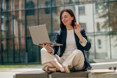 Dışarıda çalışan bir iş kadını öğrenci sanal çalışma alanında bilgisayarıyla video görüşmesi yaparken el sallıyor.