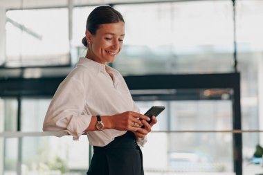 Şık giyinmiş profesyonel bir kadın akıllı telefonunu parlak bir ofiste kullanır, odaklanmasını ve nişanlanmasını arttırır.