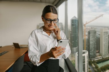 Resmi giyinmiş kendine güvenen bir iş kadını akıllı telefonunu kullanarak modern ve verimli bir çalışma alanını somutlaştırıyor.