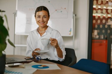 Kendine güvenen bir kadın modern bir ofiste şık masasında kahvesini yudumlarken güzel bir pozitiflik ister.