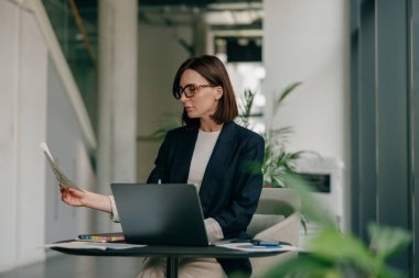Masasında dizüstü bilgisayarı ve ofisindeki bitkilerle dolu evrakları inceleyen profesyonel bir kadın.