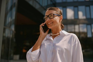 Şık gözlüklü kendine güvenen profesyonel bir kadın modern bir binanın dışında telefonla konuşurken gülümsüyor.