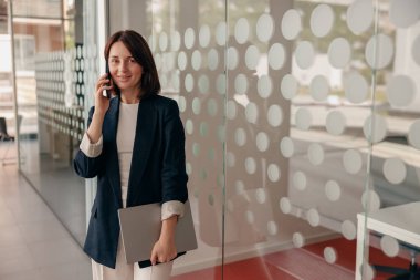 Profesyonel iş kadını modern ofiste duruyor, telefonla konuşuyor ve elinde bir dizüstü bilgisayarla güvenle gülümsüyor..