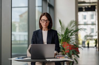 Modern ofis ortamında odaklanmış bir iş kadını, arkasında yeşil bitkiler olan bir dizüstü bilgisayar üzerinde çalışıyor..