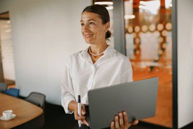 Kendine güvenen profesyonel bir kadın çağdaş bir ofis ortamında dururken elinde parlak bir laptop tutuyor.