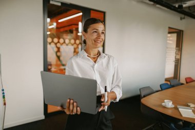 Kendine güvenen bir kadın, elinde laptopuyla şık ve modern bir ofiste duruyor. Önemli bir toplantıya hazırlanıyor.