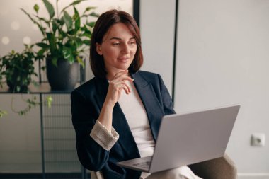 Modern bir ofiste dizüstü bilgisayar kullanan kendine güvenen bir iş kadını. Bitkileri ve dekorasyonu olan, profesyonellik saçan ve odaklanan biri.