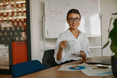 Şık gözlükler takan profesyonel bir kadın şık, modern ofis ortamında sıcak bir fincan kahve içiyor.