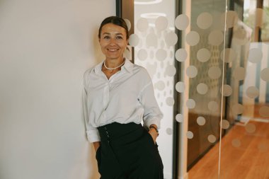 Beyaz gömlekli ve siyah pantolonlu, parlak gülüşlü bir kadın ofisinde düşünceli bir şekilde poz verir.