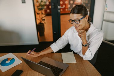 Modern bir ofis ortamında otururken özenle dizüstü bilgisayarı üzerinde çalışan kendini işine adamış ve odaklanmış profesyonel bir kadın.