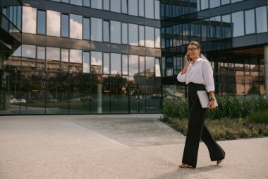 Zeki ve şık giyinmiş oldukça profesyonel bir kadın modern bir ofis alanının yanında rahatça yürüyor.