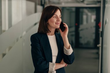 Modern bir ofis koridorunda dikilirken telefonla konuşan koyu renk ceketli kendine güvenen bir iş kadını..