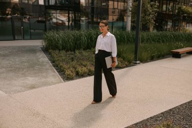 Beyaz bluz ve siyah pantolon giyen şık bir kadın modern şehir ortamında güvenle yürür..