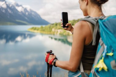 Bir yürüyüşçü akıllı telefonuyla fotoğraf çekerken nefes kesici dağlara ve sakin göle hayran olur..