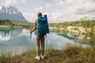 Bir yürüyüşçü heybetli dağlarla çevrili sakin bir gölün yanında duruyor, heyecan verici bir maceraya hazır.