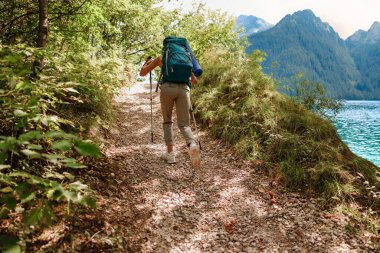 Büyük sırt çantalı bir yürüyüşçü, sakin, huzurlu bir göl ve yüksek dağların yanında güzel manzaralı bir patika boyunca yürür.