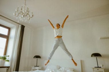 Işıkla dolu, ferah ve havadar bir yatak odasında yumuşak ve rahat bir yatakta neşeyle zıplayan bir genç.