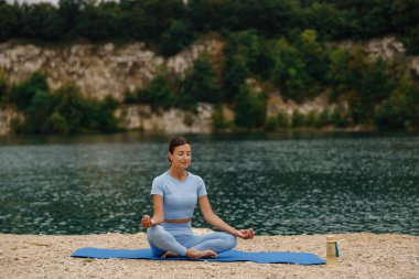 Huzurlu bir kadın, huzurlu bir gölün kenarında meditasyon yapıyor. Doğanın sağladığı sükuneti tamamen kucaklıyor.