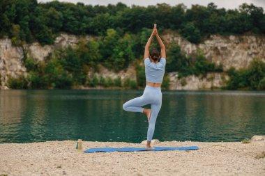 Huzur içinde yoga yapan bir insan, yemyeşil yemyeşil bir gölün yanındaki minderinin üzerinde.