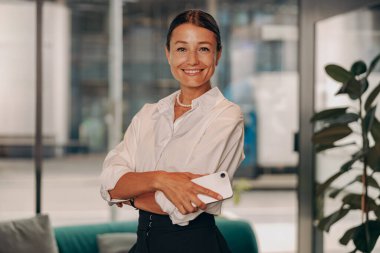 Kendine güvenen, gülümseyen bir kadın modern ofisinde gururla telefonunu tutuyor ve başarısını sergiliyor.