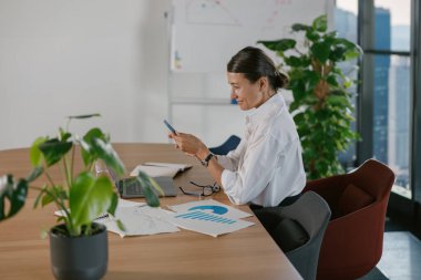 Modern ofis ortamında, yetenekli profesyonel bir kadın akıllı telefonunu çalışırken etkin bir şekilde kullanır.