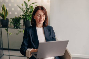 Profesyonel bir kadın, modern bir ofis alanında dizüstü bilgisayar kullanırken gülümsüyor. Bitkiler ve şık dekorlarla.