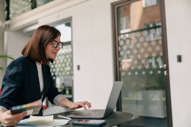 Profesyonel bir kadın, modern ofis ortamında kitap ve belgelerle çevrili bir dizüstü bilgisayar kullanıyor.