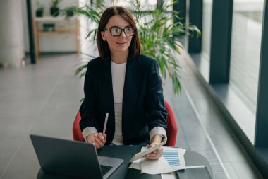 Bir iş kadını, çağdaş bir ofis ortamında dizüstü bilgisayar ve belgeler kullanarak çalışmaktadır.