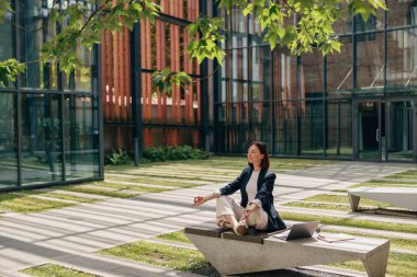 Bir kadın, açık havada modern bir binanın önündeki bankta oturmuş huzur dolu bir anın tadını çıkarıyor.