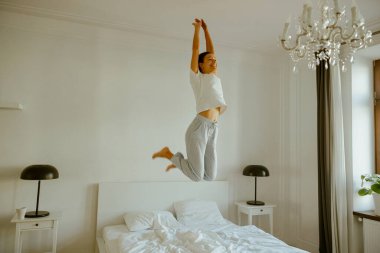 Bir insan mutlu bir şekilde yumuşak bir yatakta zıplıyor, parlak bir yatak odasında saf mutluluğun özünü yakalıyor.