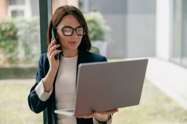 Profesyonel iş kadını, telefon ve dizüstü bilgisayarda birden fazla iş yapıyor, modern ofis ortamında çalışıyor..