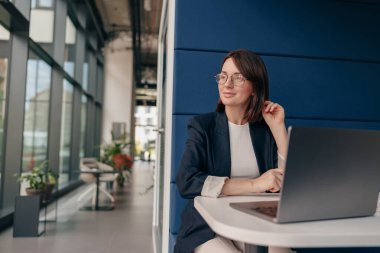 Ofiste dizüstü bilgisayarlı profesyonel bir kadın, dışarıya bakıyor, düşünceli bir ifade, çağdaş bir dekor..