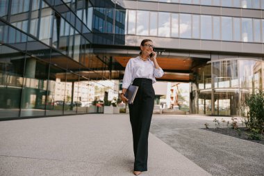 Modern bir şehirde telefon görüşmesi yapan profesyonel bir kadın güvenli bir şekilde dışarı çıkıyor.