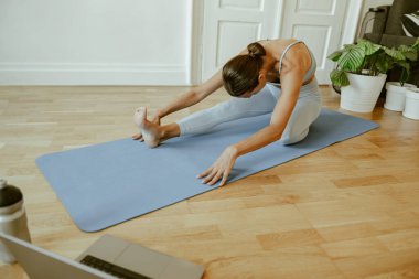 Bir kadın evdeki minderinde yoga esneme hareketi yapıyor, sağlığın ve farkındalığın önemini vurguluyor.